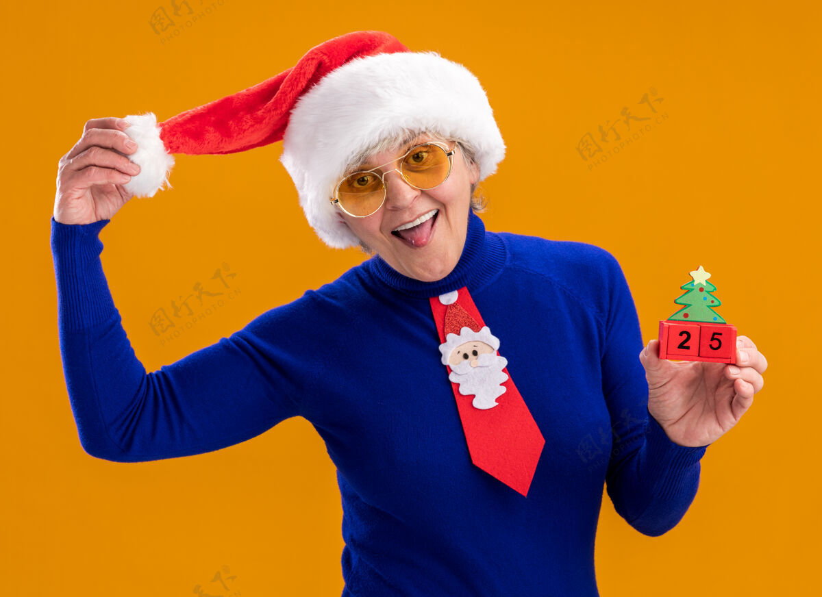 老人戴着太阳眼镜 戴着圣诞帽 系着圣诞领带的快乐的老妇人伸出舌头 把圣诞树装饰物孤立地放在橙色的背景上 并留有复制空间快乐舌头圣诞树
