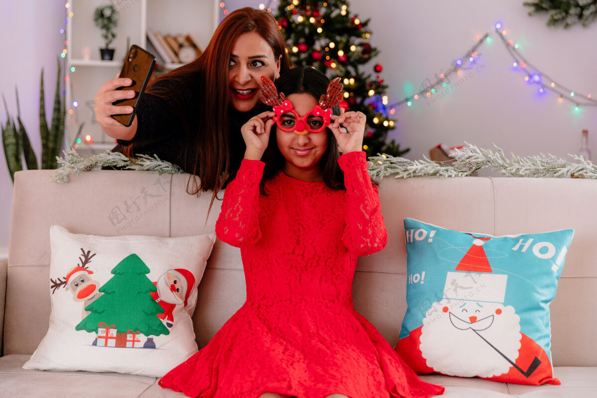 快乐微笑的母亲拍下了女儿戴着驯鹿眼镜坐在沙发上在家里享受圣诞节时光的照片微笑女儿家