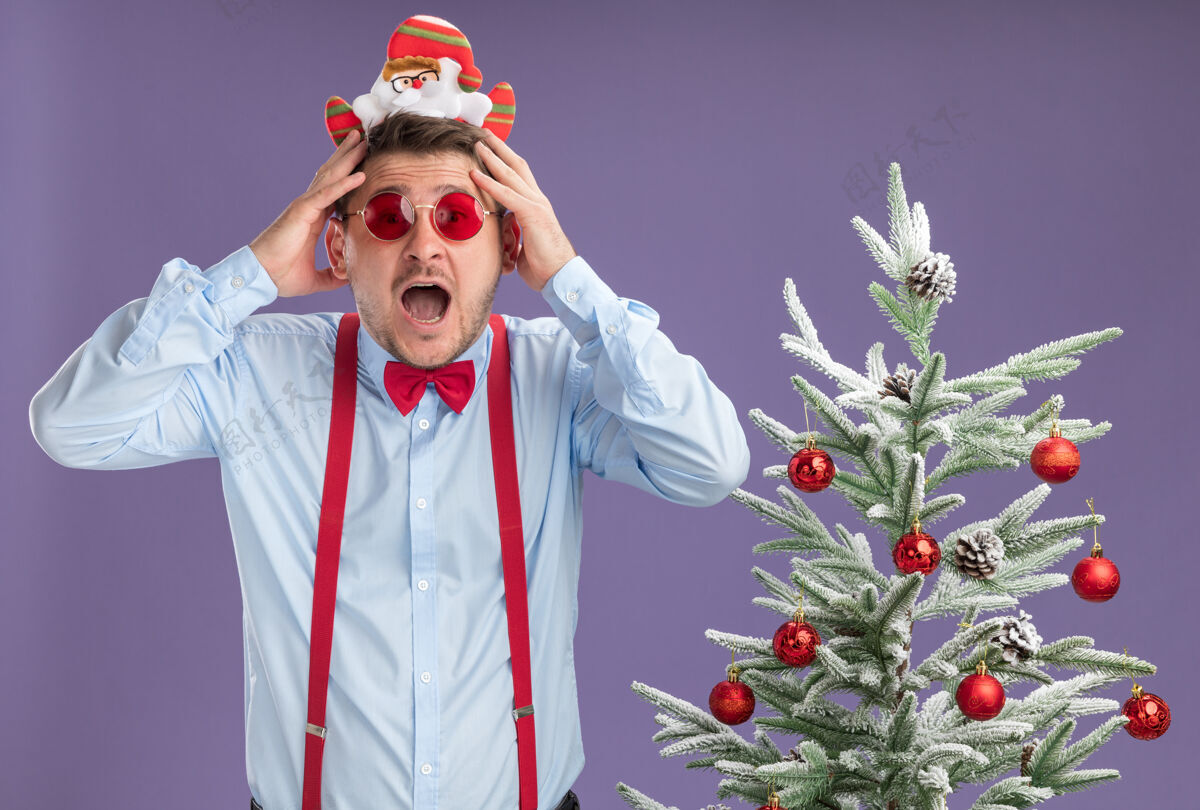 领带站在圣诞树旁边的一个戴着圣诞老人和红眼镜 系着吊带领结的年轻人 手放在紫色的墙上 惊讶又惊讶吊带圣诞节轮辋