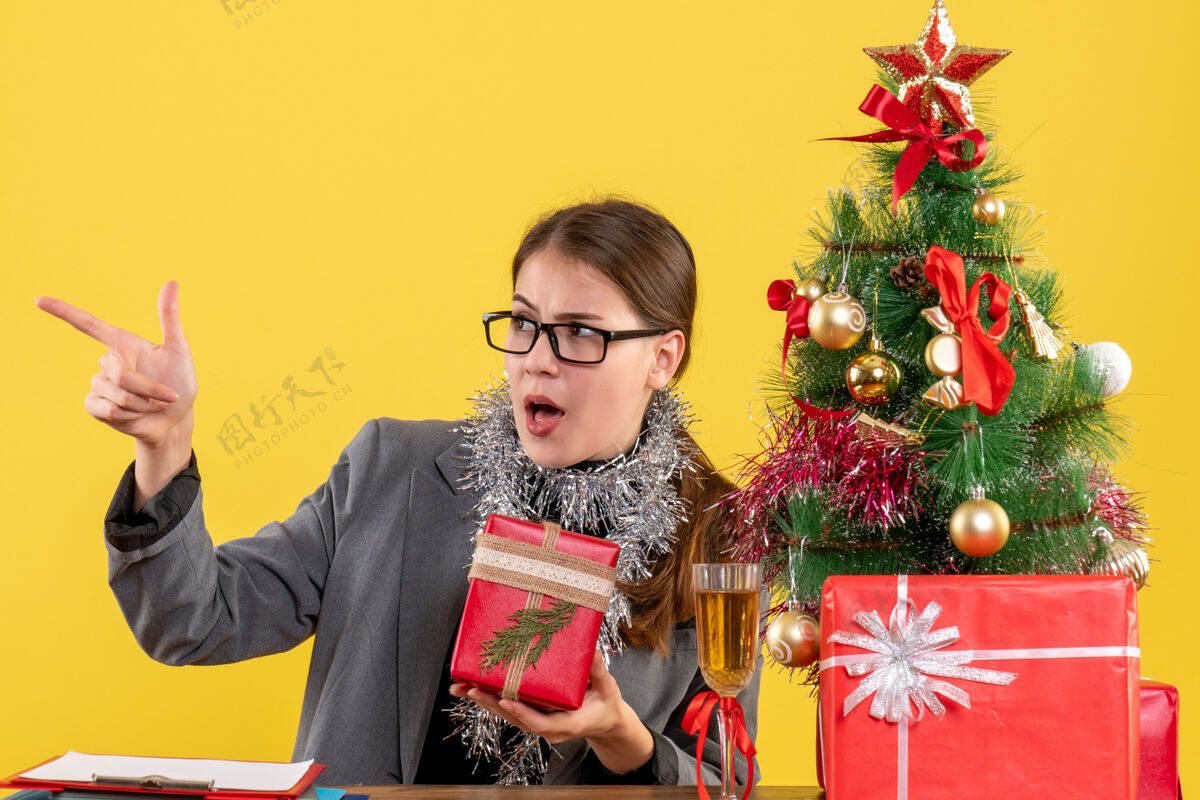 鸡尾酒前视图漫步的女孩戴着眼镜坐在桌边看着左边的圣诞树和鸡尾酒礼物看微笑树