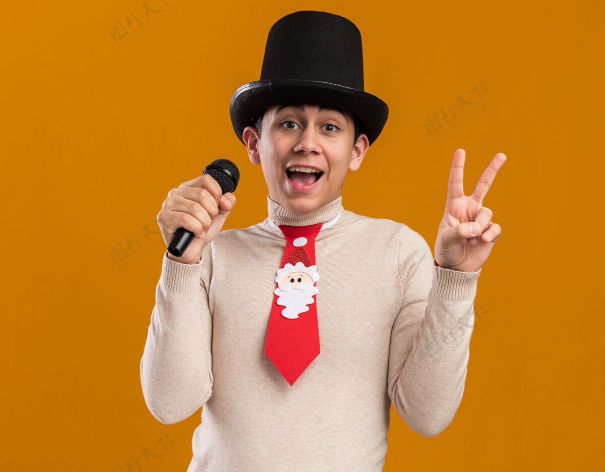 快乐戴着圣诞领带戴着帽子的年轻人在麦克风上发表讲话 在黄色的墙上显示出与世隔绝的和平姿态穿着圣诞节和平
