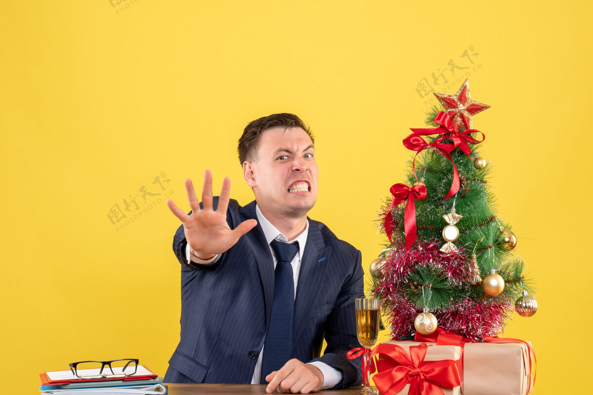 前面愤怒的男人在圣诞树旁的桌子前做停车标志 黄色的墙上摆着礼物行政人员树男