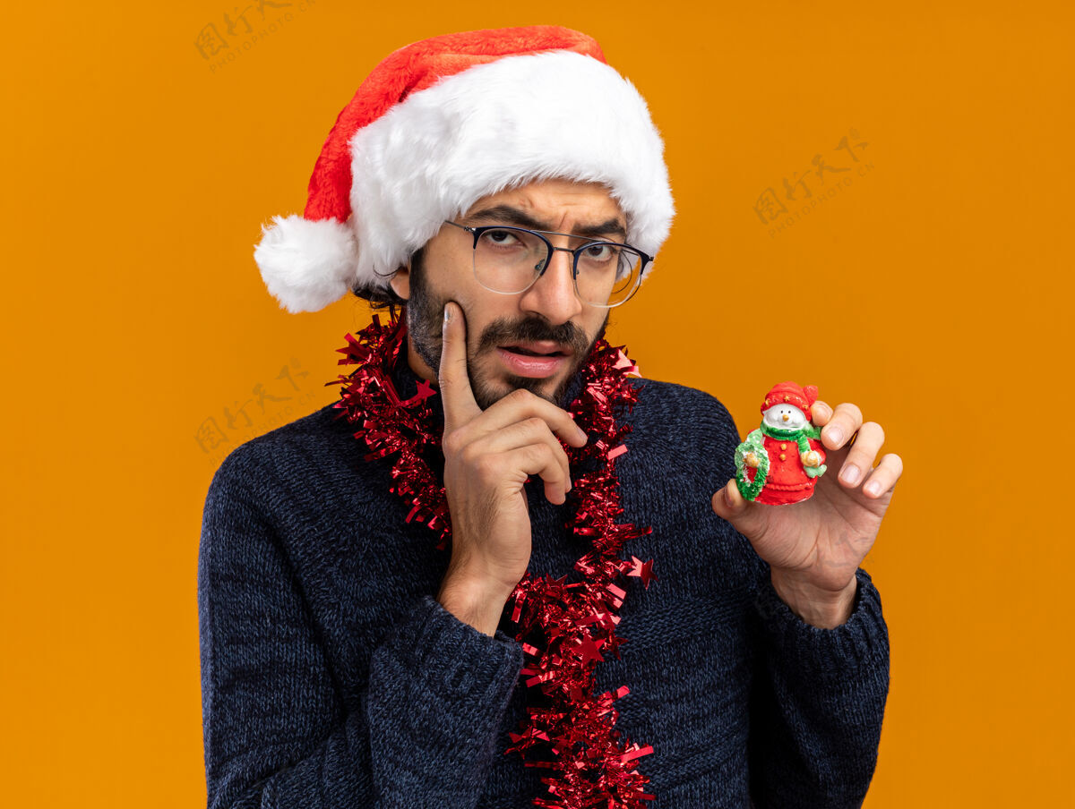 脸颊想着年轻帅哥戴着圣诞帽脖子上戴着花环拿着玩具把手指放在脸颊上孤立在橙色背景上思想手指抱