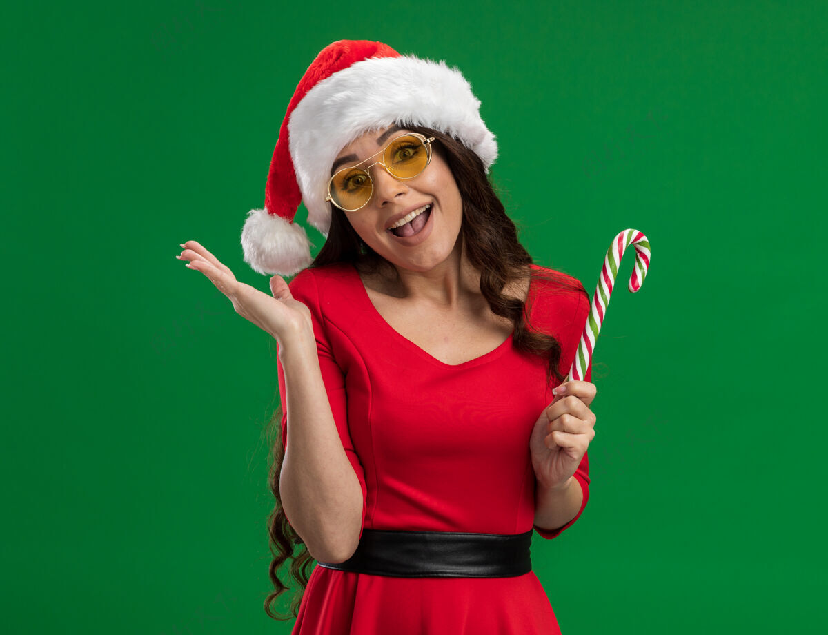 圣诞老人快乐的年轻漂亮女孩戴着圣诞帽戴着圣诞眼镜拿着圣诞糖果手杖看着相机显示空手孤立在绿色背景上帽子持有眼镜