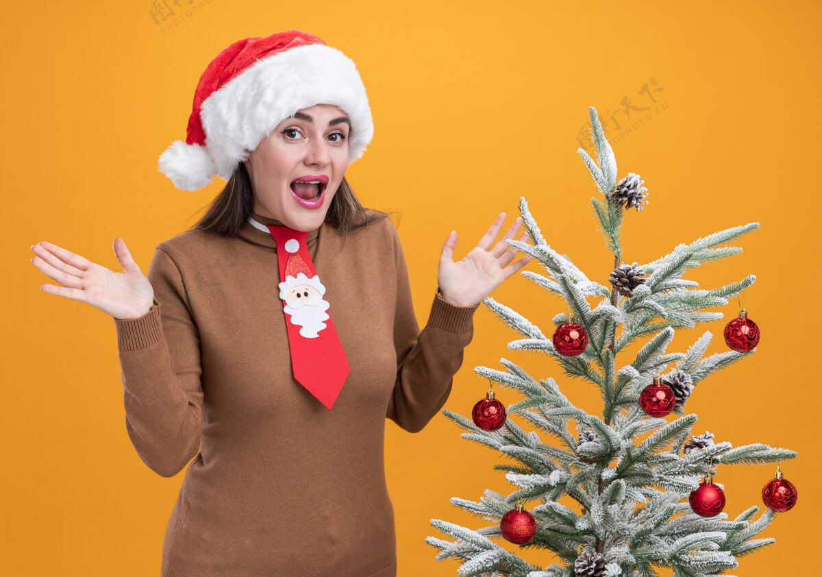 年轻兴奋的年轻漂亮女孩戴着圣诞帽打着领带站在圣诞树旁摊开双手孤立在橙色的墙上附近站着传播