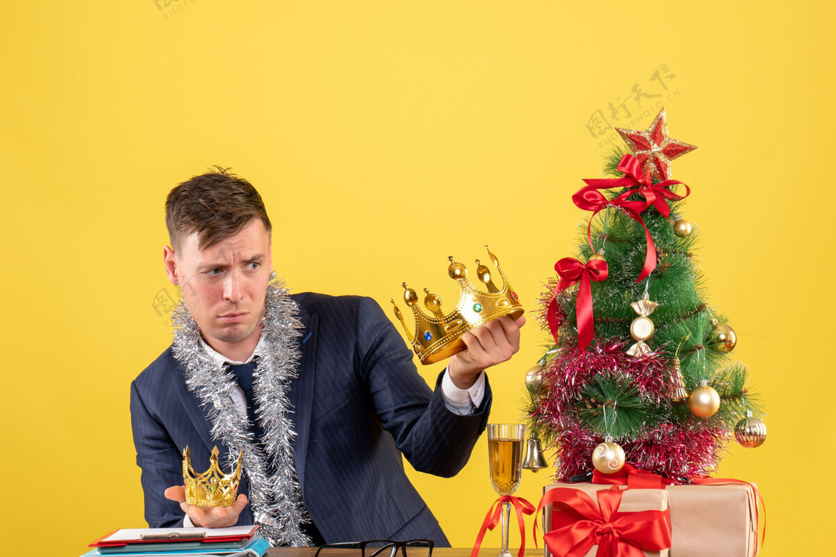 西装商人坐在圣诞树旁的桌子旁比较他的王冠和黄色墙上的礼物的正面图礼物树人