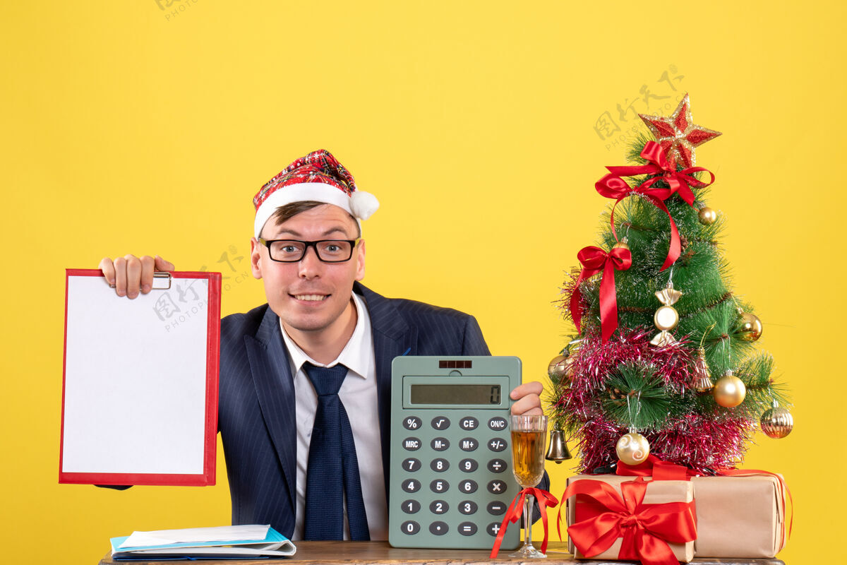 坐圣诞树旁的桌子上坐着一个戴着圣诞帽的笑脸男人 黄色的墙上挂着礼物桌子商人商务