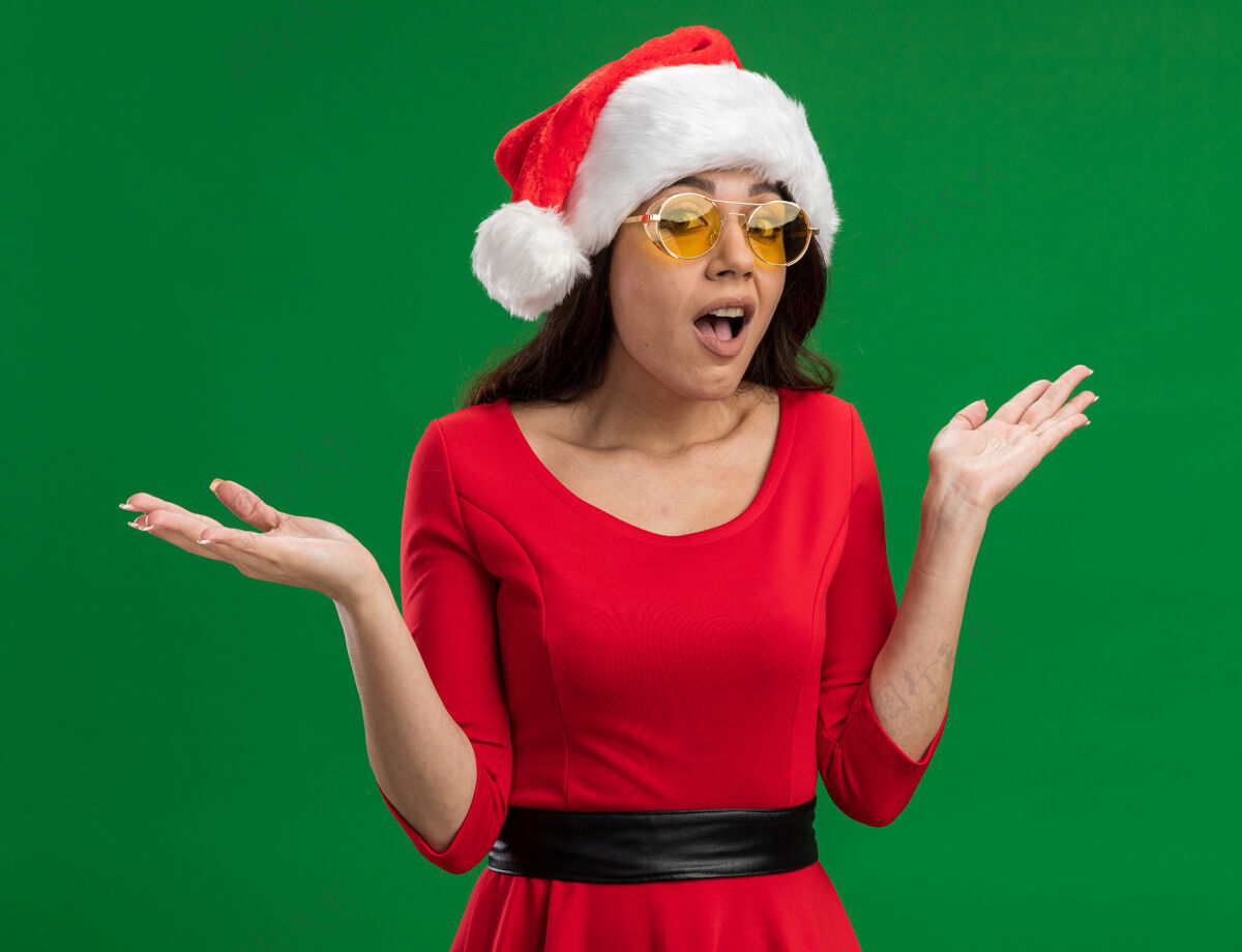 手无知的年轻漂亮女孩戴着圣诞帽 戴着眼镜 两手空空地看着隔离在绿色背景上的相机圣诞老人空虚眼镜