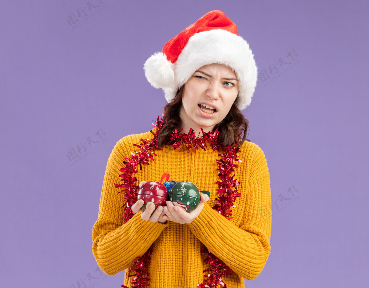 恼怒恼怒的年轻斯拉夫女孩戴着圣诞帽 脖子上戴着花环 手里拿着玻璃球饰物 看着紫色背景上孤立的一面 还有复制空间周围年轻斯拉夫