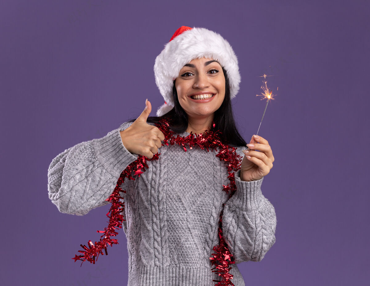 穿着快乐的年轻白人女孩戴着圣诞帽 脖子上戴着金箔花环 手里拿着节日火花灯 孤立地在紫色的墙上竖起大拇指圣诞节周围显示