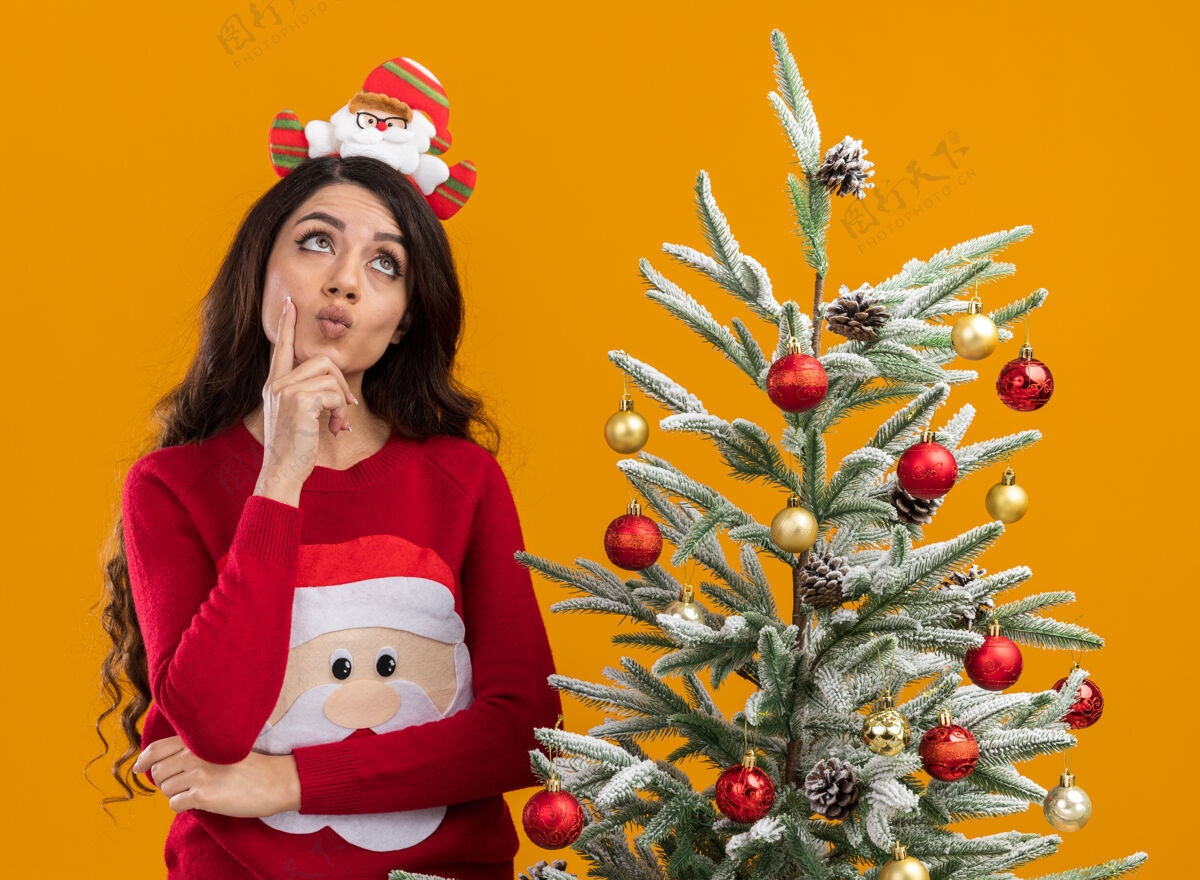 圣诞体贴的年轻漂亮女孩 戴着圣诞老人的头带和毛衣 站在装饰好的圣诞树旁 手放在下巴上 在橙色的背景下孤立地抬头看下巴橙色圣诞快乐