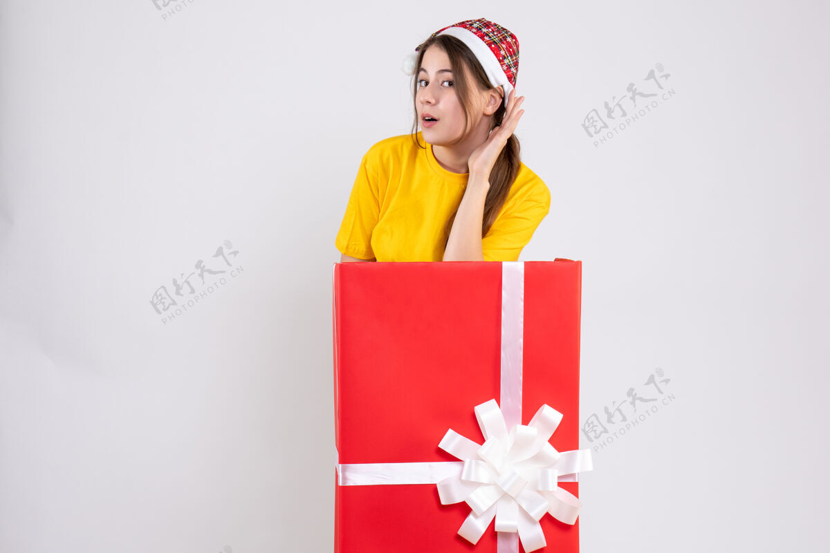 举行戴着圣诞帽的窥探女孩站在白色大圣诞礼物后面爱国者购物者圣诞节