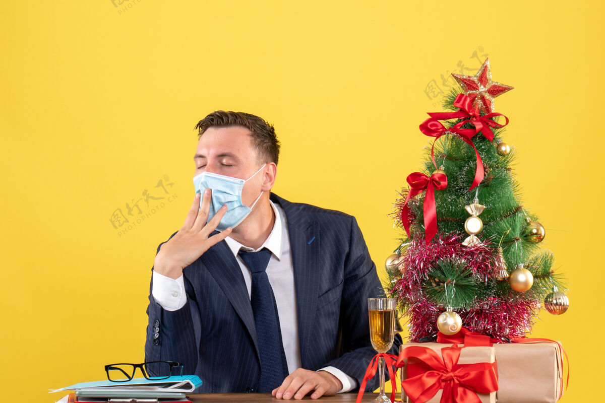 成人昏昏欲睡的商人坐在圣诞树旁的桌子前 黄色的礼物商务人专业