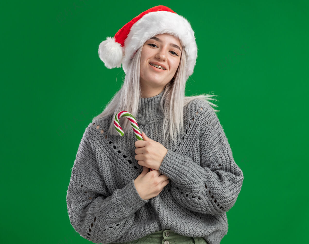 金发年轻的金发女郎穿着冬衣 戴着圣诞帽 手里拿着糖果手杖 站在绿色的背景下 开心而积极地微笑着看着镜头快乐手杖圣诞