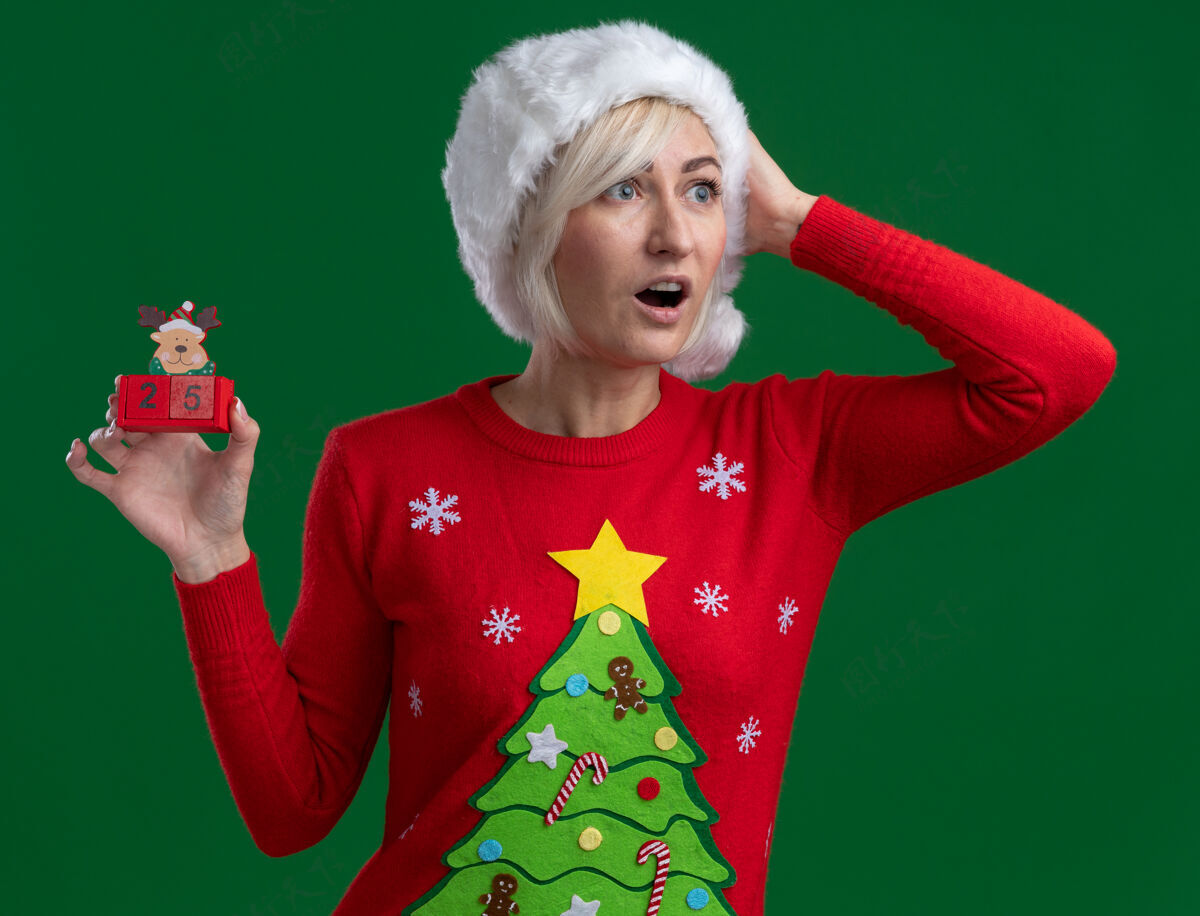 穿令人印象深刻的中年金发女人戴着圣诞帽和毛衣拿着圣诞驯鹿玩具和日期看一边保持手放在头上隔离在绿色墙上金发女人印象深刻
