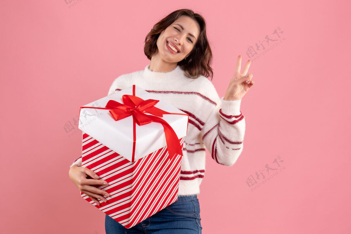 正面带着圣诞礼物的年轻女子在粉色墙上微笑的正面图人模特礼物