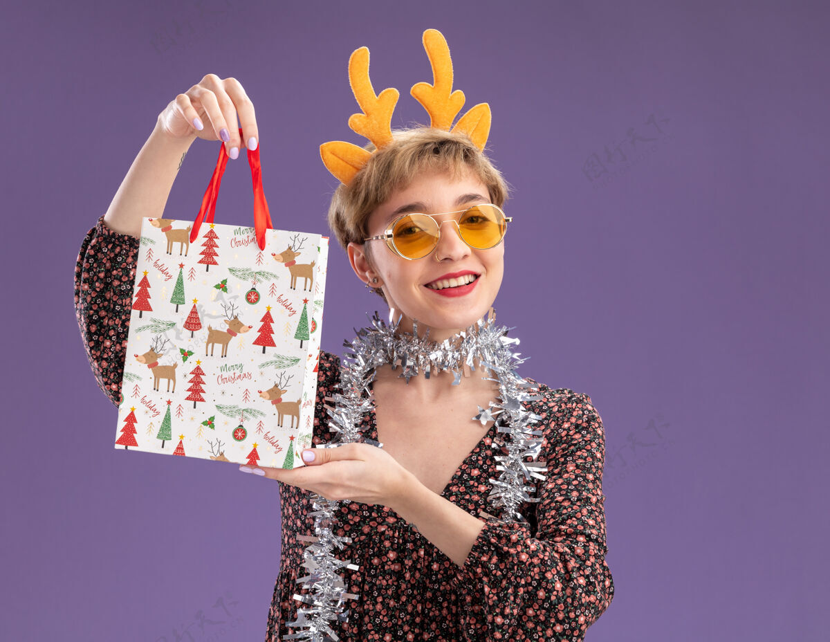 漂亮快乐的年轻漂亮女孩戴着驯鹿鹿角头带 脖子上戴着金箔花环 戴着眼镜 手里拿着圣诞礼包 隔离在紫色的墙上 留着复印空间快乐年轻围着