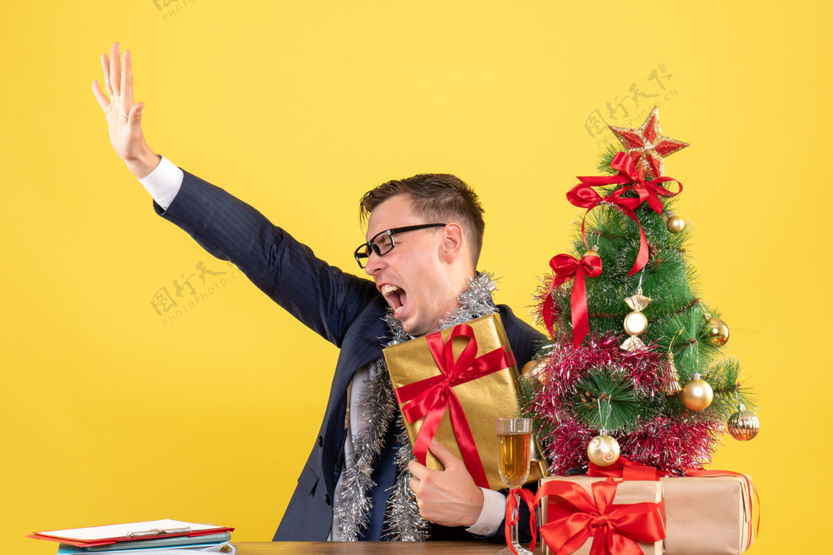 前面愤怒的男人张开手坐在圣诞树旁的桌子前 黄色的墙上摆着礼物肖像微笑人