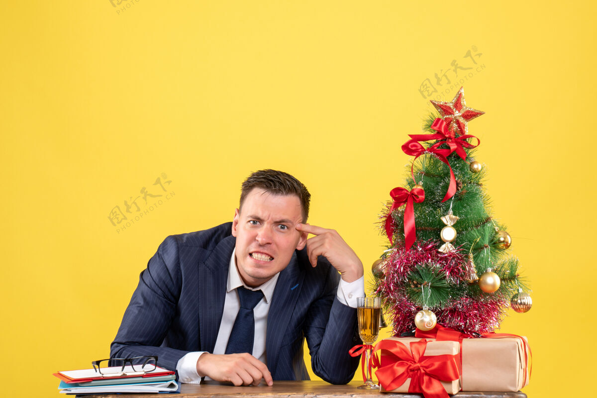 男性正面图愤怒的年轻人坐在圣诞树旁的桌子旁 黄色墙壁上免费赠送礼物西装礼物圣诞节