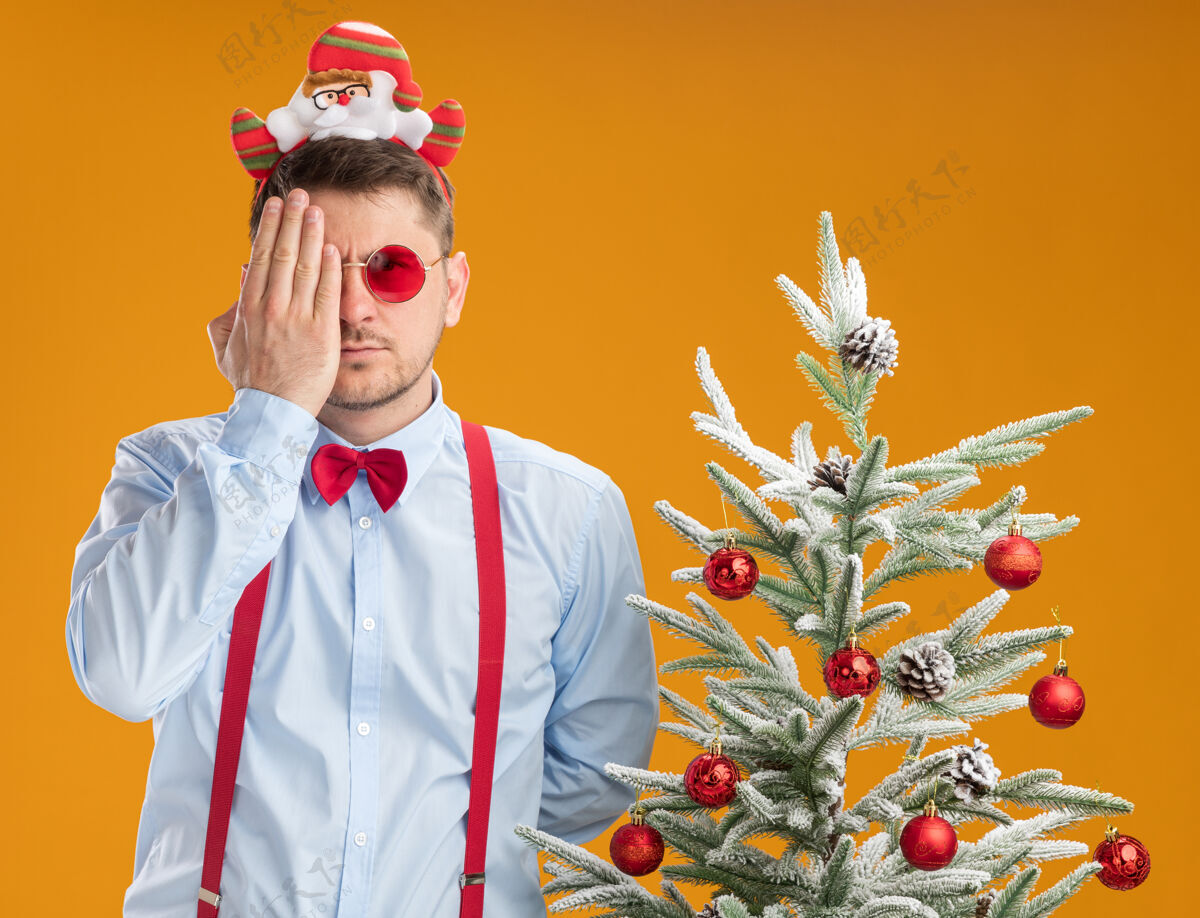 领带戴着吊带领结的年轻人站在圣诞树旁 戴着圣诞老人和红眼镜 用手遮住一只眼睛 看着橙色背景下的摄像机圣诞手蝴蝶结
