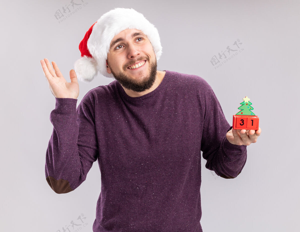 毛衣身穿紫色毛衣 头戴圣诞帽的快乐快乐的年轻人站在白色背景上 展示着新年约会的方块 脸上洋溢着欢快的笑容年高兴年轻