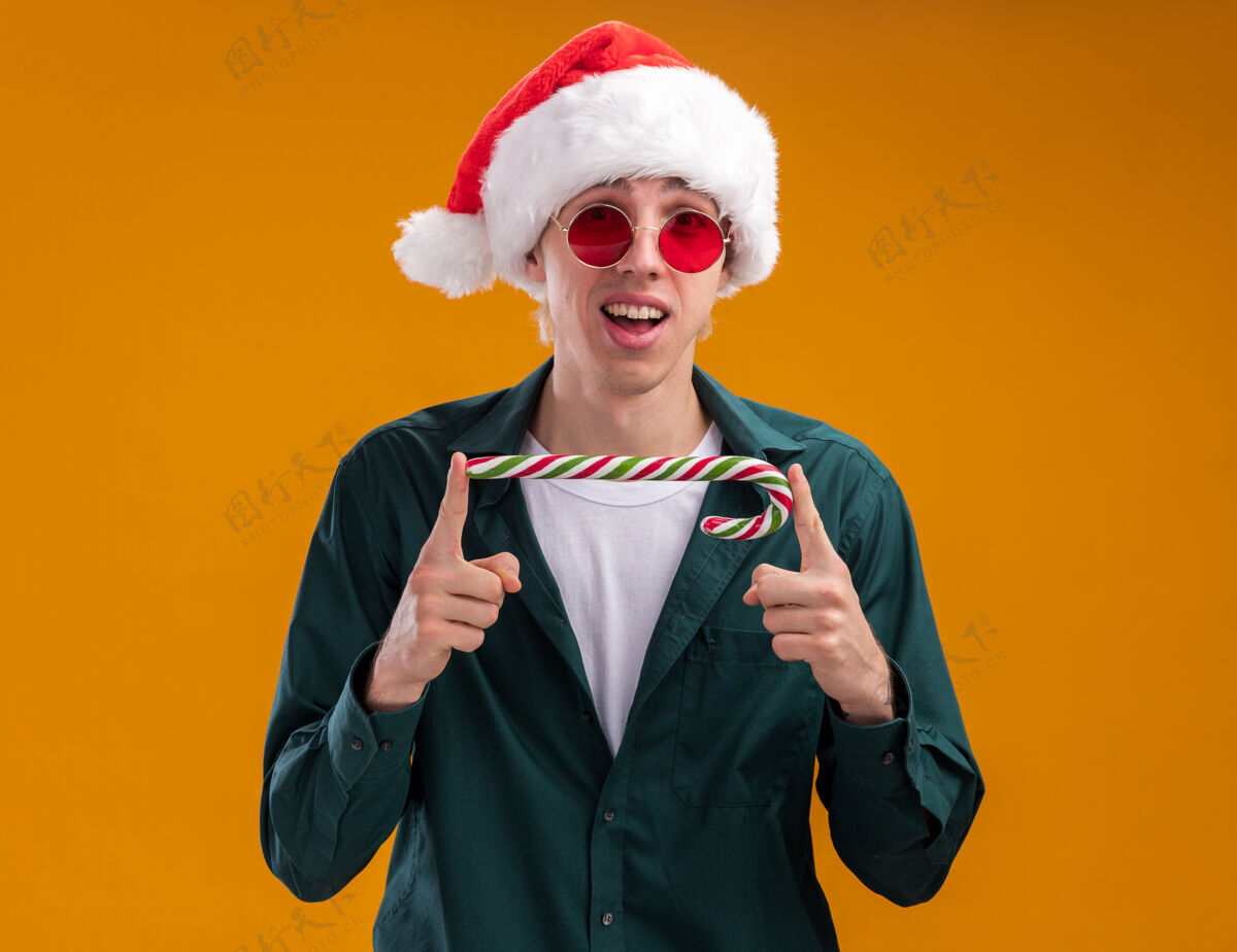手杖令人印象深刻的年轻金发男子戴着圣诞帽和眼镜手持圣诞甜手杖水平看着相机孤立的橙色背景甜蜜年轻印象深刻