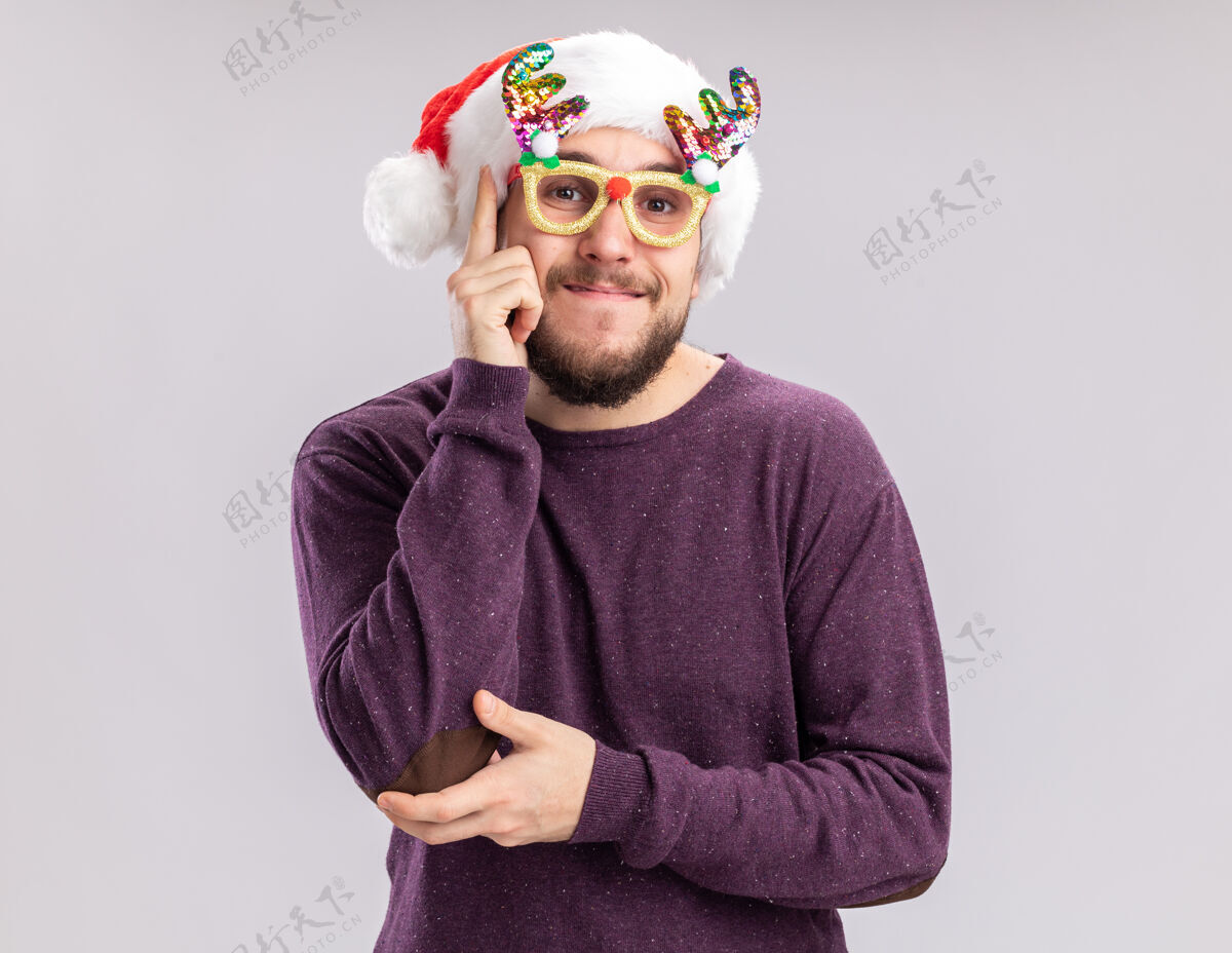 圣诞老人一个穿着紫色毛衣 戴着圣诞帽 戴着滑稽眼镜的快乐的年轻人站在白色的背景下看着镜头 开心地微笑着站着搞笑年轻