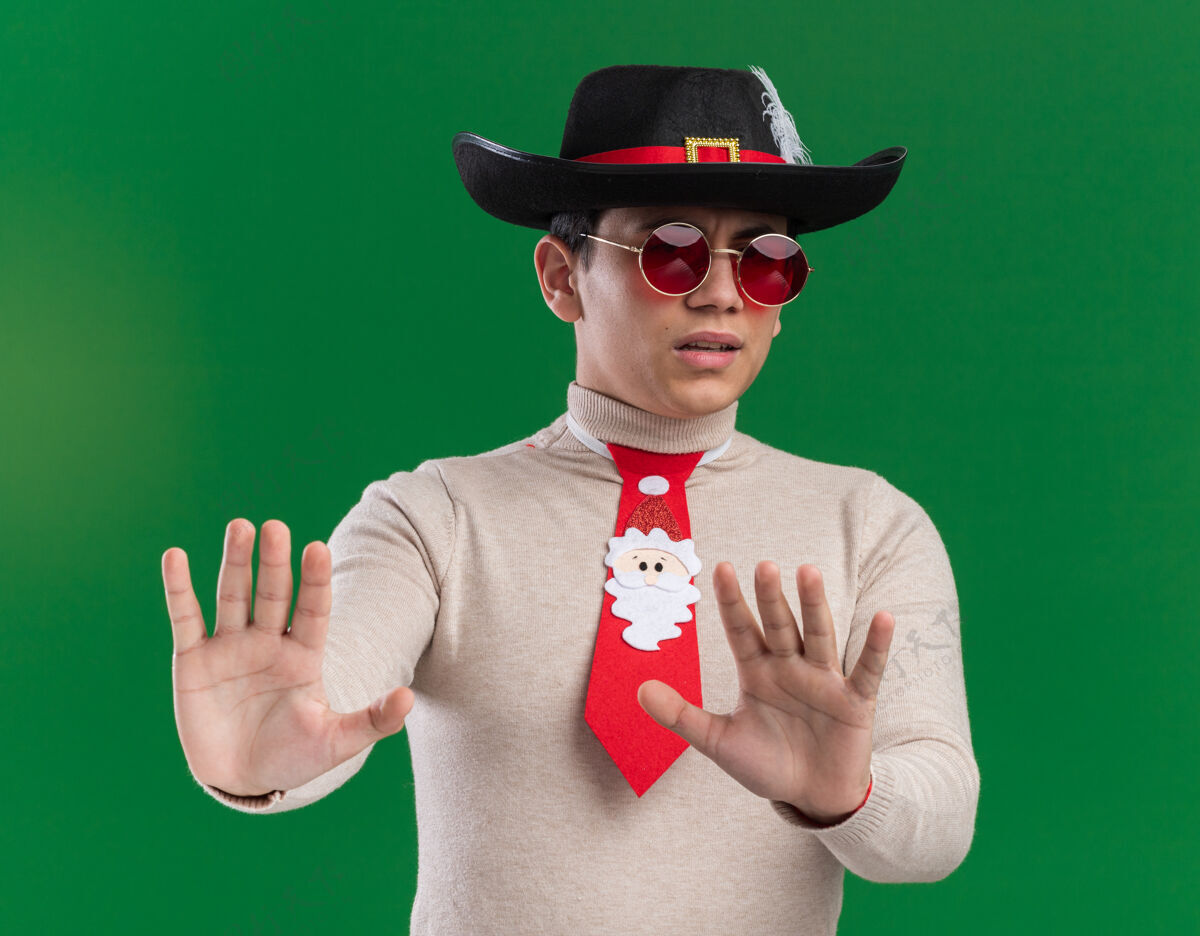 手势一个戴着圣诞领带帽子 戴着眼镜 在绿色背景上显示停止手势的年轻人吓坏了年轻圣诞节帽子