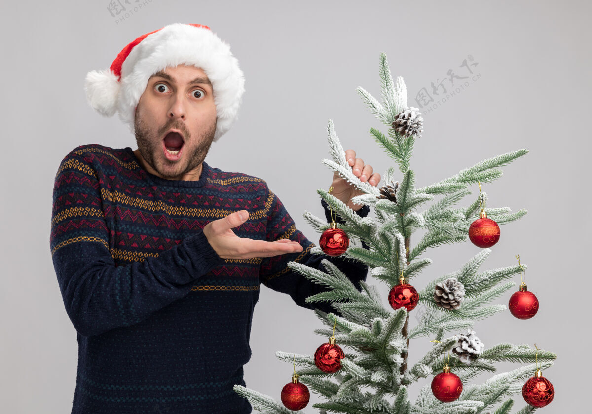 震惊震惊的年轻白种人戴着圣诞帽站在圣诞树旁抚摸着它看着隔离在白色背景下的相机相机圣诞树