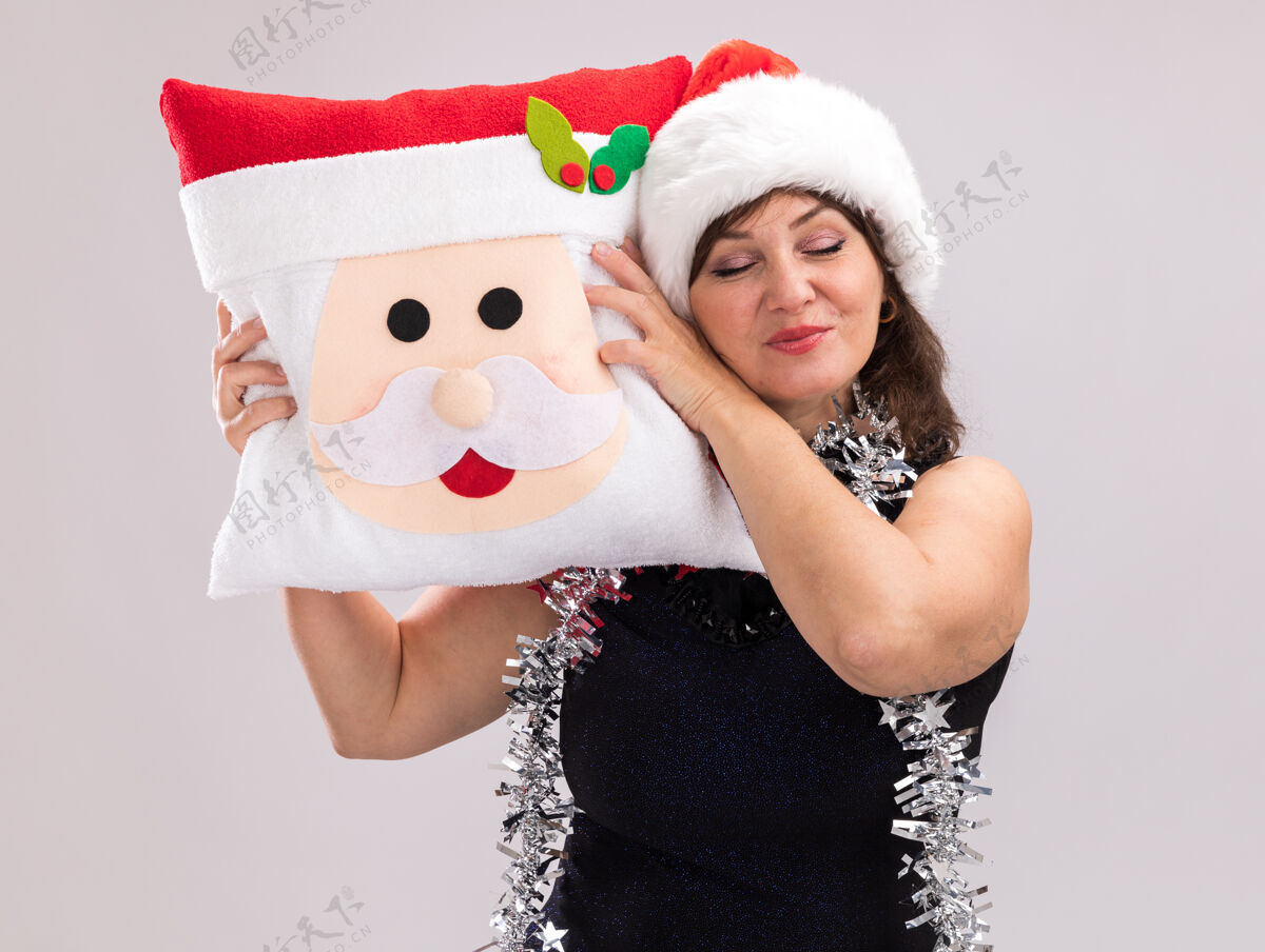 脖子高兴的中年妇女戴着圣诞帽 脖子上戴着金箔花环 抱着圣诞老人的枕头 用它抚摸着头 闭着眼睛隔离在白色的背景上高兴眼睛花环