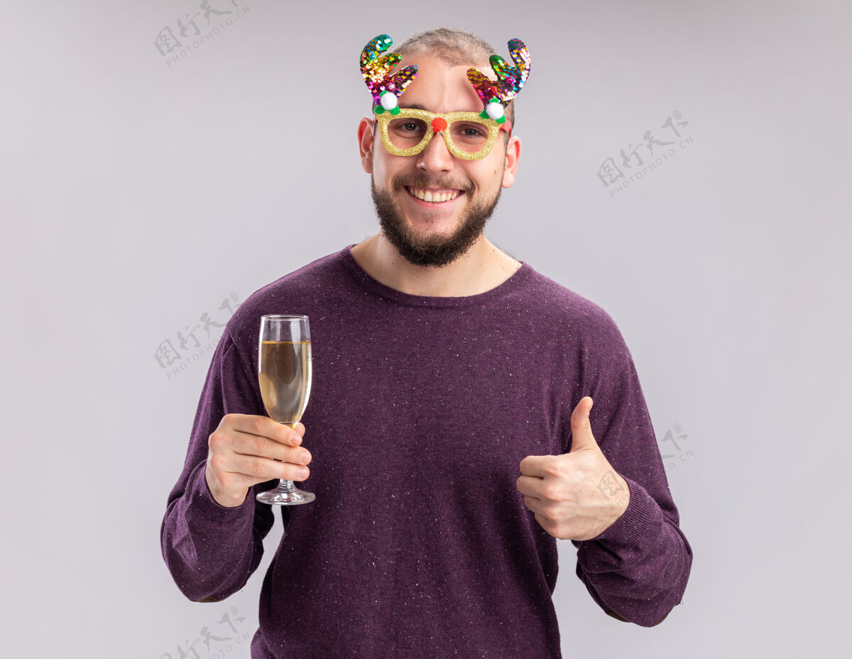 眼镜一个穿着紫色毛衣戴着滑稽眼镜的快乐的年轻人 手里拿着一杯香槟 面带微笑地看着相机 脚上穿鞋 竖起大拇指站在白色的背景上香槟玻璃杯穿鞋