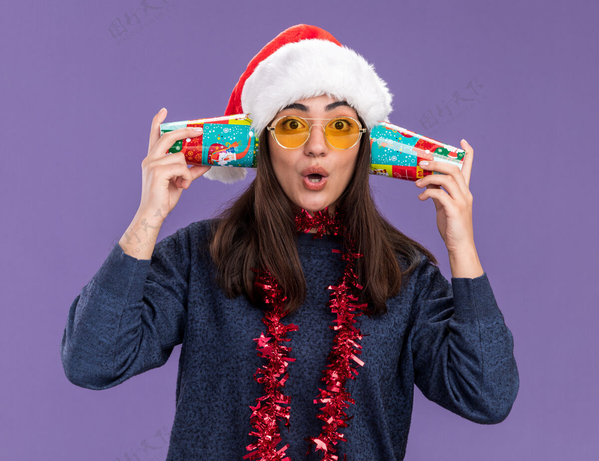 杯子戴着太阳眼镜 戴着圣诞帽 脖子上戴着花环的年轻白种人女孩惊讶地把纸杯举在耳朵旁 紫色背景上 留有复印空间女孩脖子举行