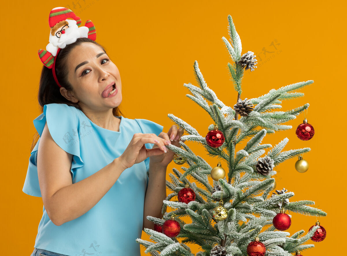 积极身着蓝色上衣的快乐年轻女子戴着有趣的圣诞环装饰圣诞树快乐而积极地伸出舌头站在橙色的背景上快乐圣诞树