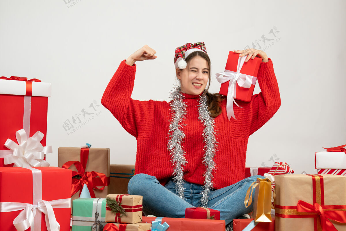 手势戴着圣诞帽的吓坏了的女孩在白色的礼物旁边摆着获胜的姿势胜利礼物害怕