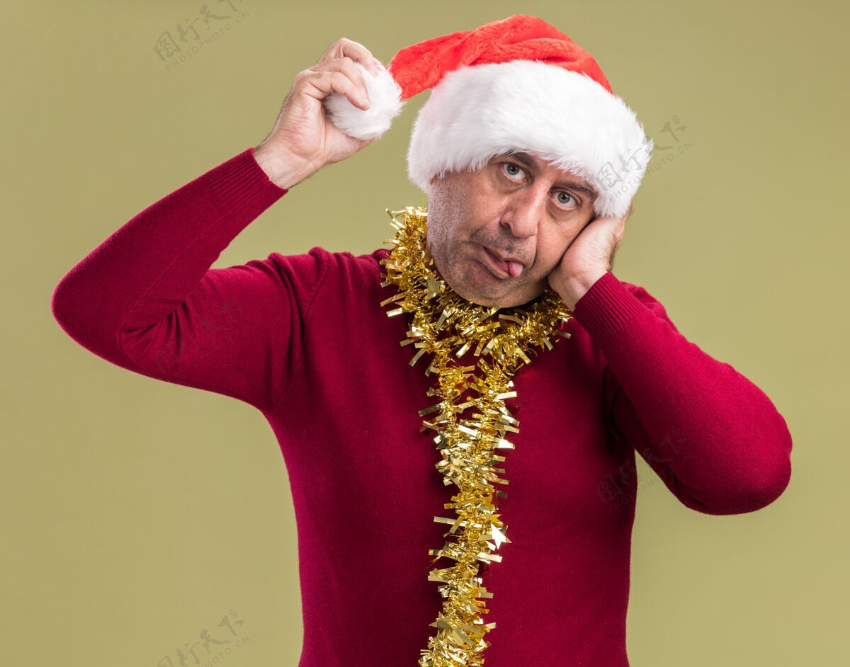 中间快乐的中年男子戴着圣诞老人的帽子 脖子上戴着金属丝 看着相机触摸着他的帽子 伸出舌头站在绿色的背景上周围快乐站立