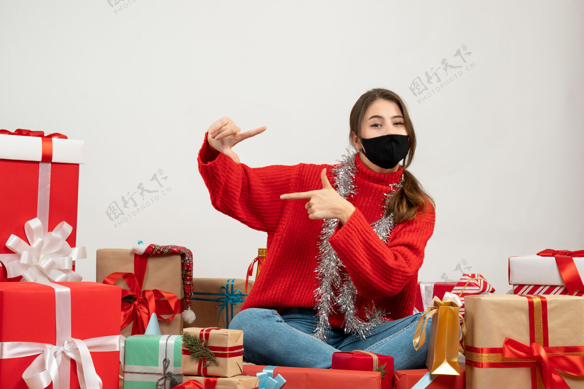 坐着穿着红色毛衣 戴着黑色面具的年轻女孩正在制作相机标志 围坐在白色的礼物上礼物帽子礼物