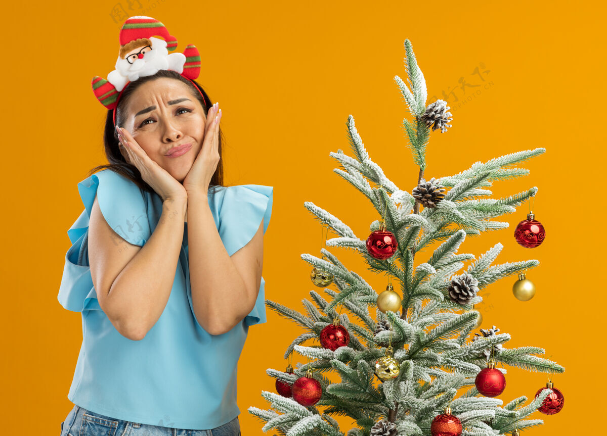 边缘身穿蓝色上衣的年轻女子头戴滑稽的圣诞戒指 抬头困惑地看着站在橙色背景下的圣诞树旁的双手树橙色有趣