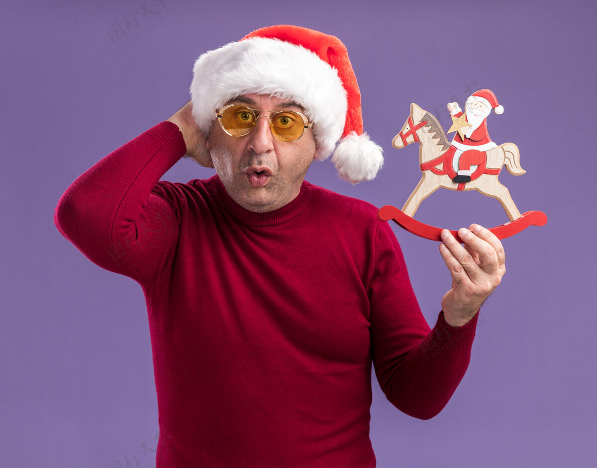 站着中年男子戴着圣诞帽 戴着黄色眼镜 手里拿着圣诞玩具 站在紫色的背景下惊讶地看着相机圣诞老人年龄眼镜