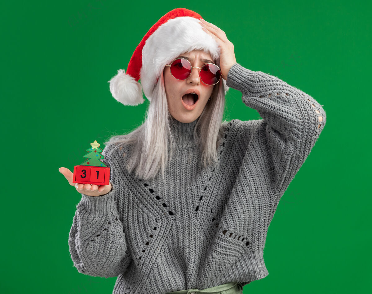 举行穿着冬季毛衣 戴着圣诞老人帽 戴着红眼镜 手里拿着玩具立方体 新年快乐 手放在头上 站在绿色背景上 看起来既困惑又担心手头站着