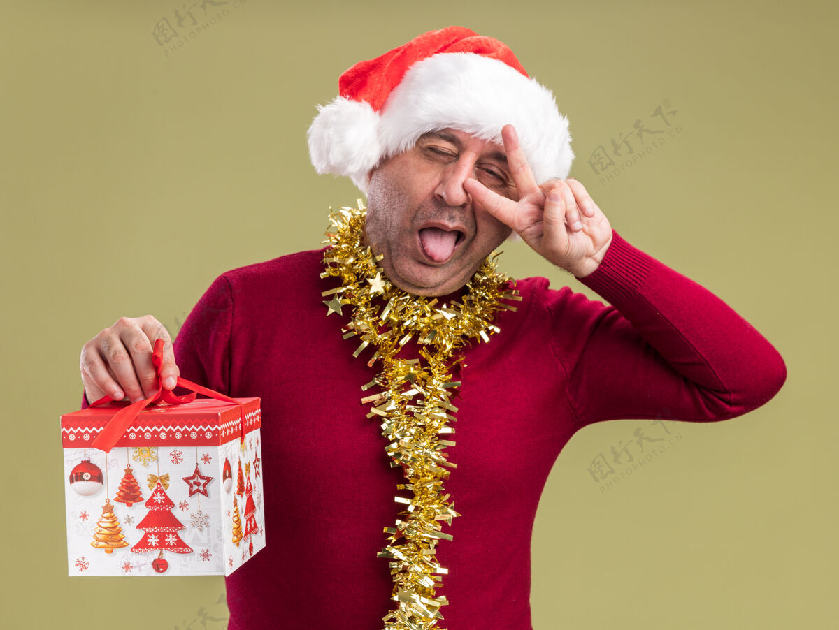 圣诞节快乐快乐的中年男子戴着圣诞老人帽 脖子上戴着金属丝 手里拿着圣诞礼物 看着伸出舌头的相机 绿色背景上立着v字标志站立年龄礼物