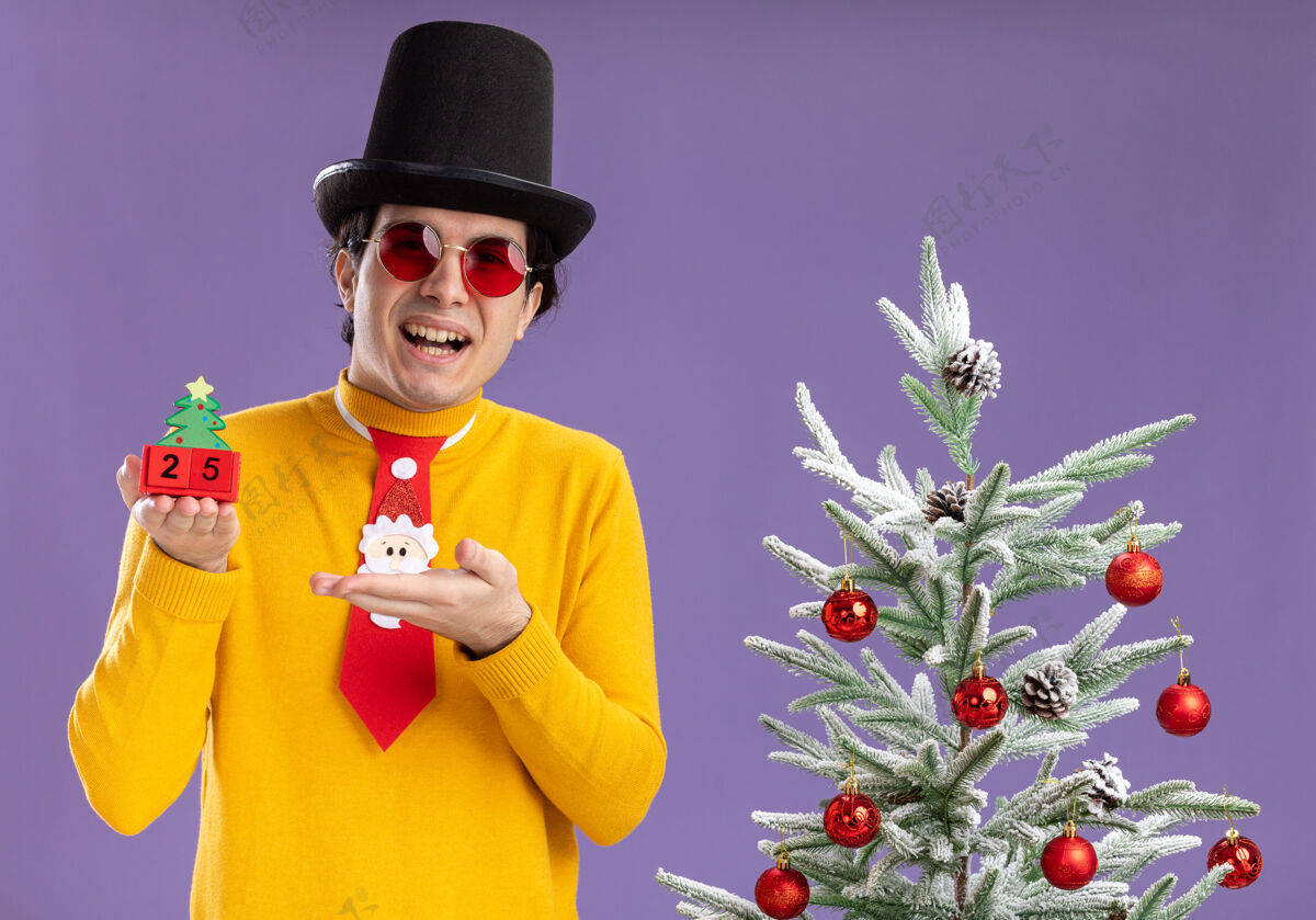 快乐身穿黄色高领毛衣 戴着眼镜 戴着黑色帽子 打着滑稽领带的快乐年轻人手持25号立方体 手挽手微笑着站在圣诞树旁手臂帽子二十