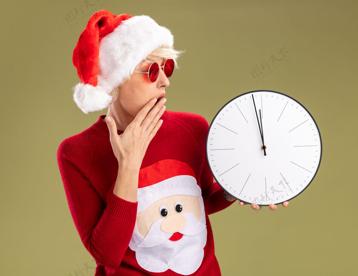 保管关心的金发女郎戴着圣诞帽 穿着圣诞老人的圣诞毛衣 戴着眼镜 手放在嘴上 看着橄榄绿墙上孤立的时钟年轻圣诞老人橄榄