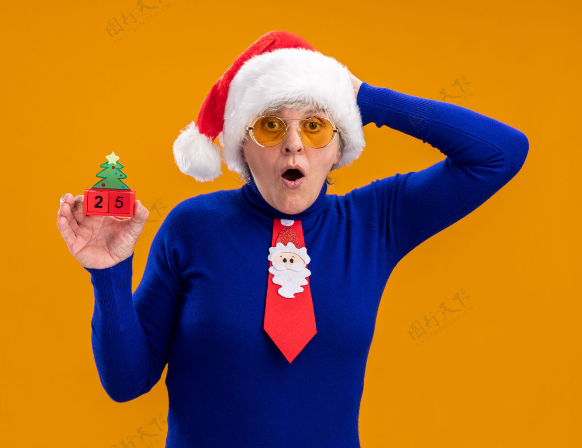 举行戴着太阳眼镜 戴着圣诞帽 打着圣诞领带的震惊的老妇人拿着圣诞树饰物 把手放在孤立的橙色背景上 留有复制空间眼镜帽子装饰品