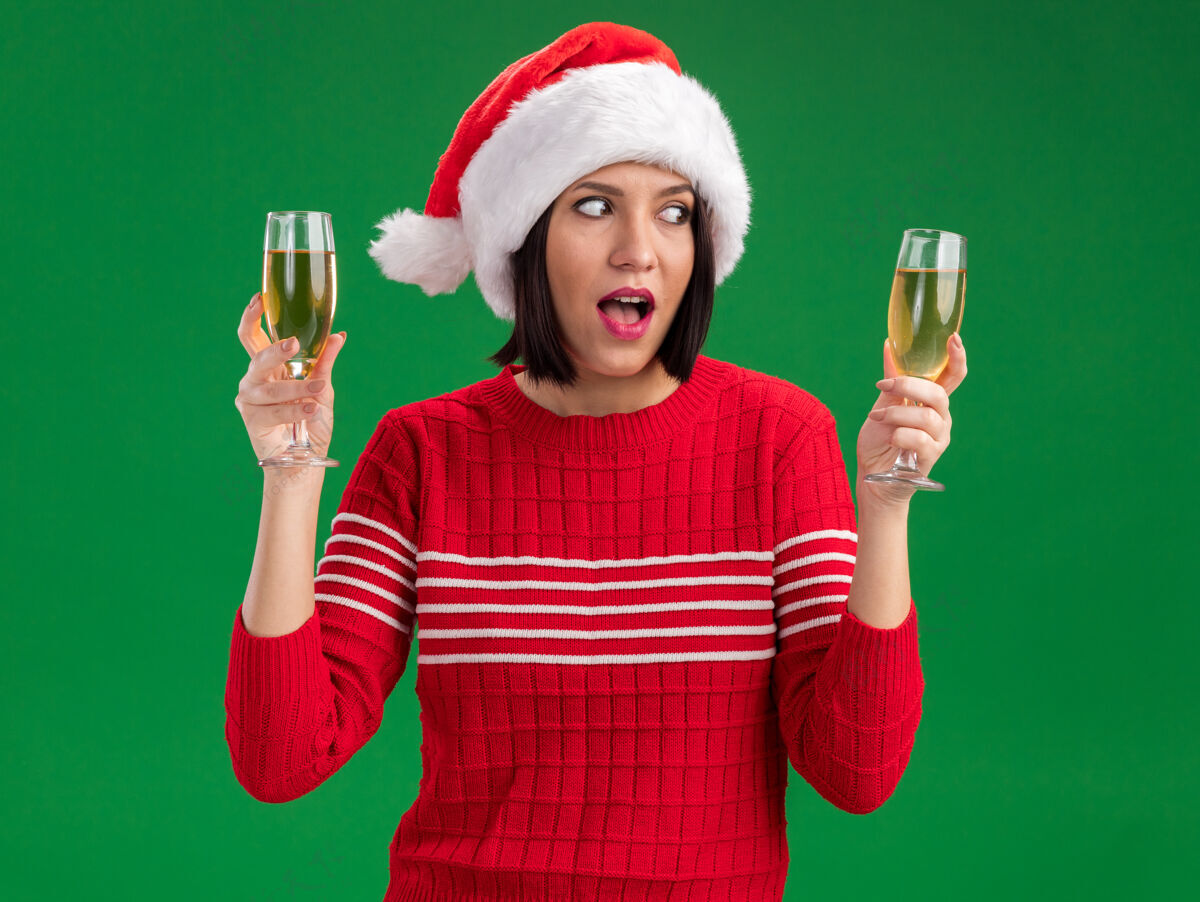 圣诞老人印象深刻的年轻女孩戴着圣诞帽拿着两杯香槟看着一个孤立的绿色背景年轻人香槟眼镜