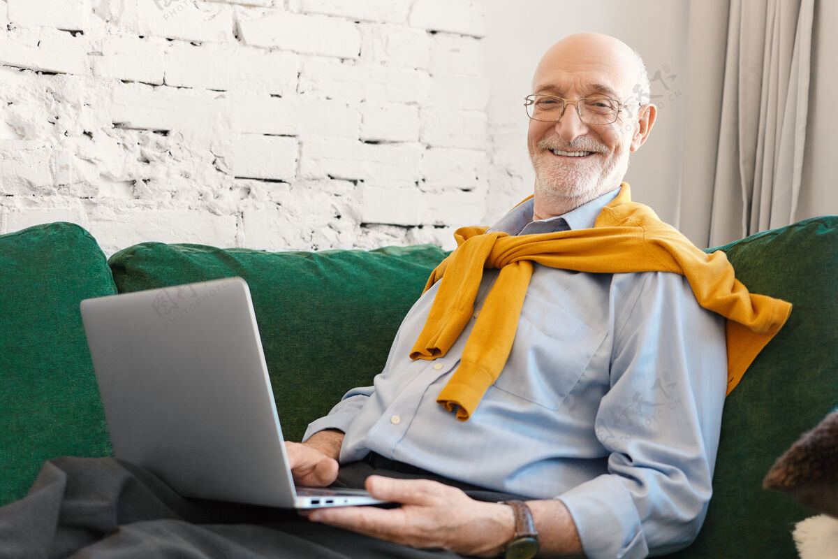 互联网戴着眼镜 穿着优雅的衣服 坐在沙发上 拿着笔记本电脑 对着镜头微笑 为在线报纸或博客撰写商业文章的正面有吸引力的男记者的室内照片房间60岁房子