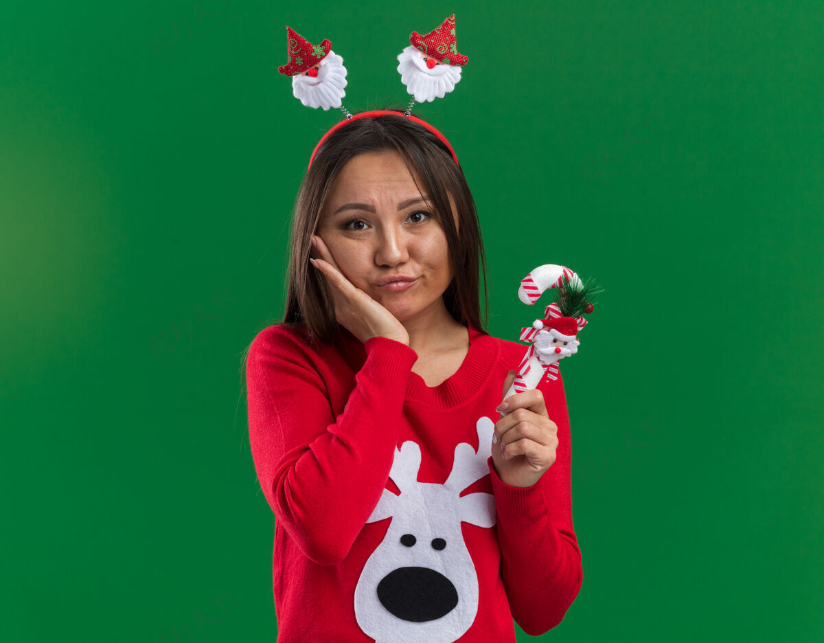 脸颊悲伤的年轻亚洲女孩戴着圣诞发箍和毛衣拿着圣诞糖果把手放在脸颊上隔离在绿色背景上亚洲人悲伤手