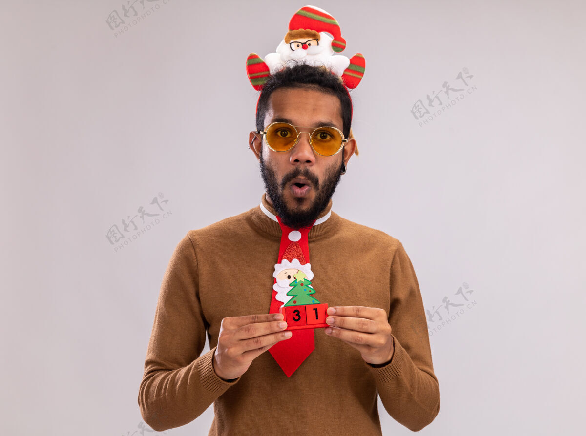 毛衣一个穿着棕色毛衣 头戴圣诞老人戒指 打着有趣的红色领带的非裔美国人拿着新年约会玩具立方体 站在白色背景下惊讶地看着相机圣诞老人头日期