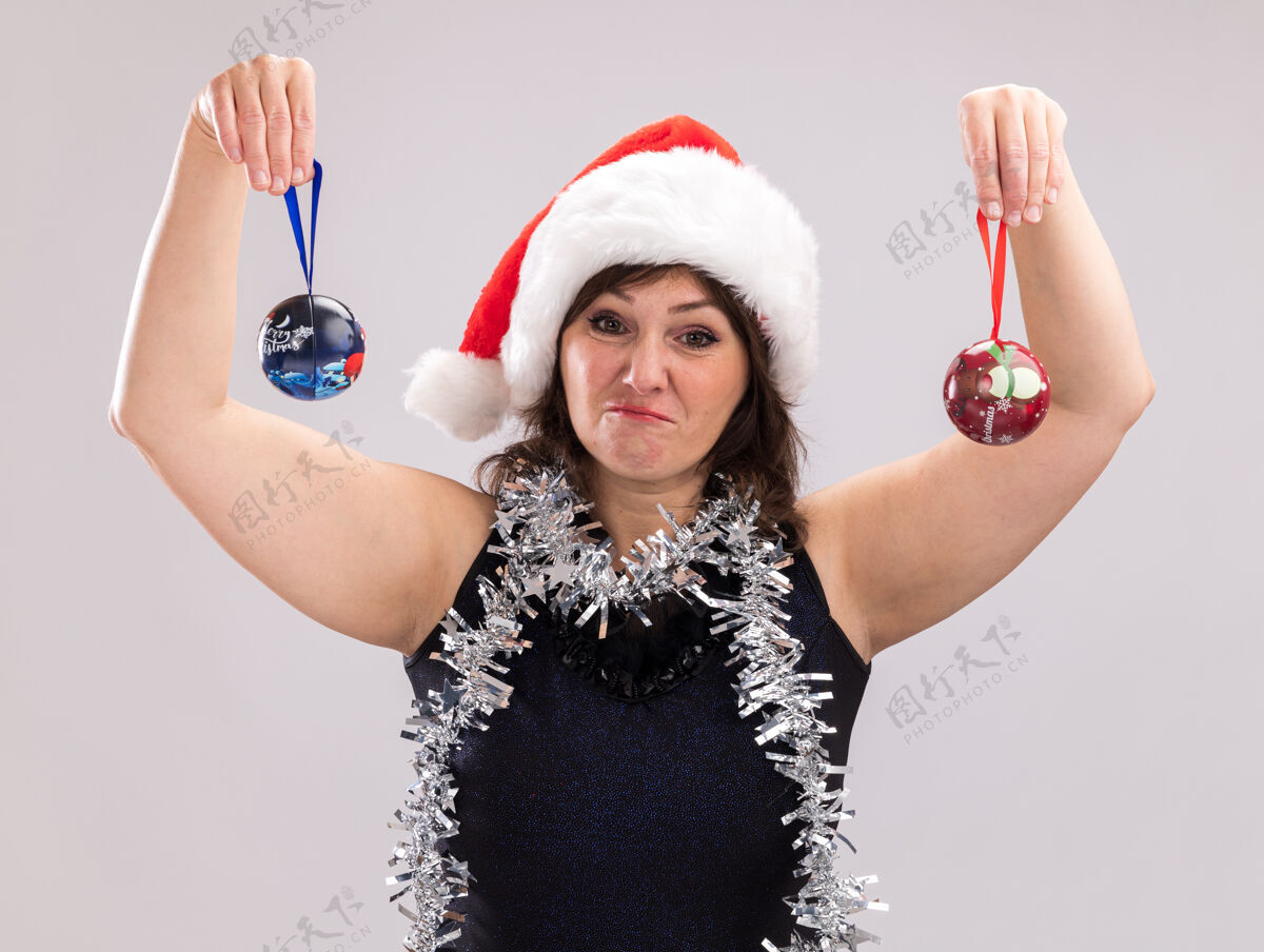 围着笨手笨脚的中年妇女戴着圣诞帽 脖子上戴着金属丝花环 举起圣诞饰品 看着隔离在白色背景上的相机中年人圣诞饰品脖子