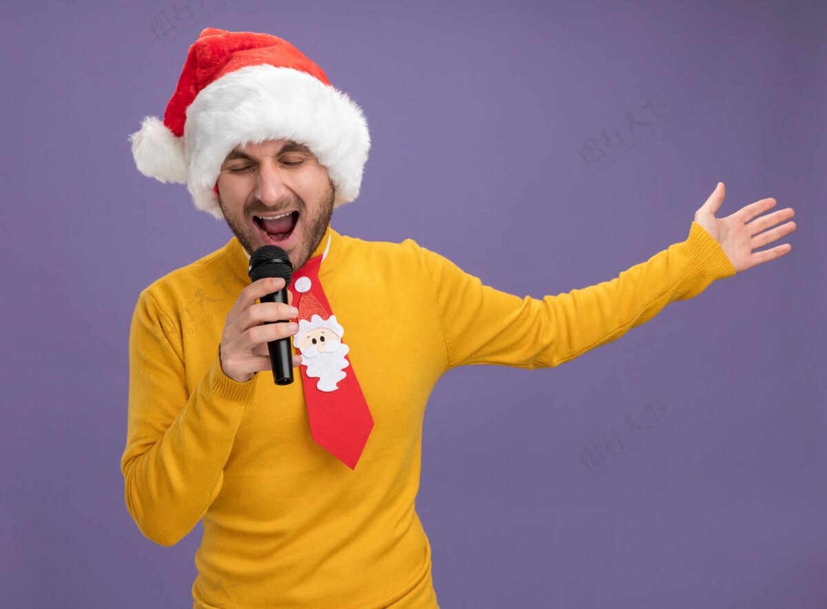 圣诞节戴着圣诞帽打着领带的白人年轻人举着麦克风 闭着眼睛 伸出手 在紫色背景上孤立地唱歌帽子闭上年轻