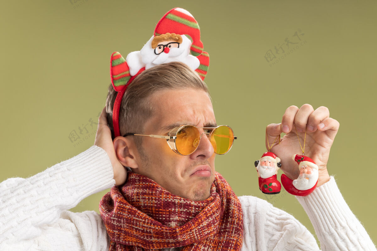 圣诞老人迷茫的年轻帅哥戴着圣诞老人的头带和围巾 拿着圣诞老人看圣诞饰品 把手放在脑后孤立在橄榄绿的墙上头头带迷茫